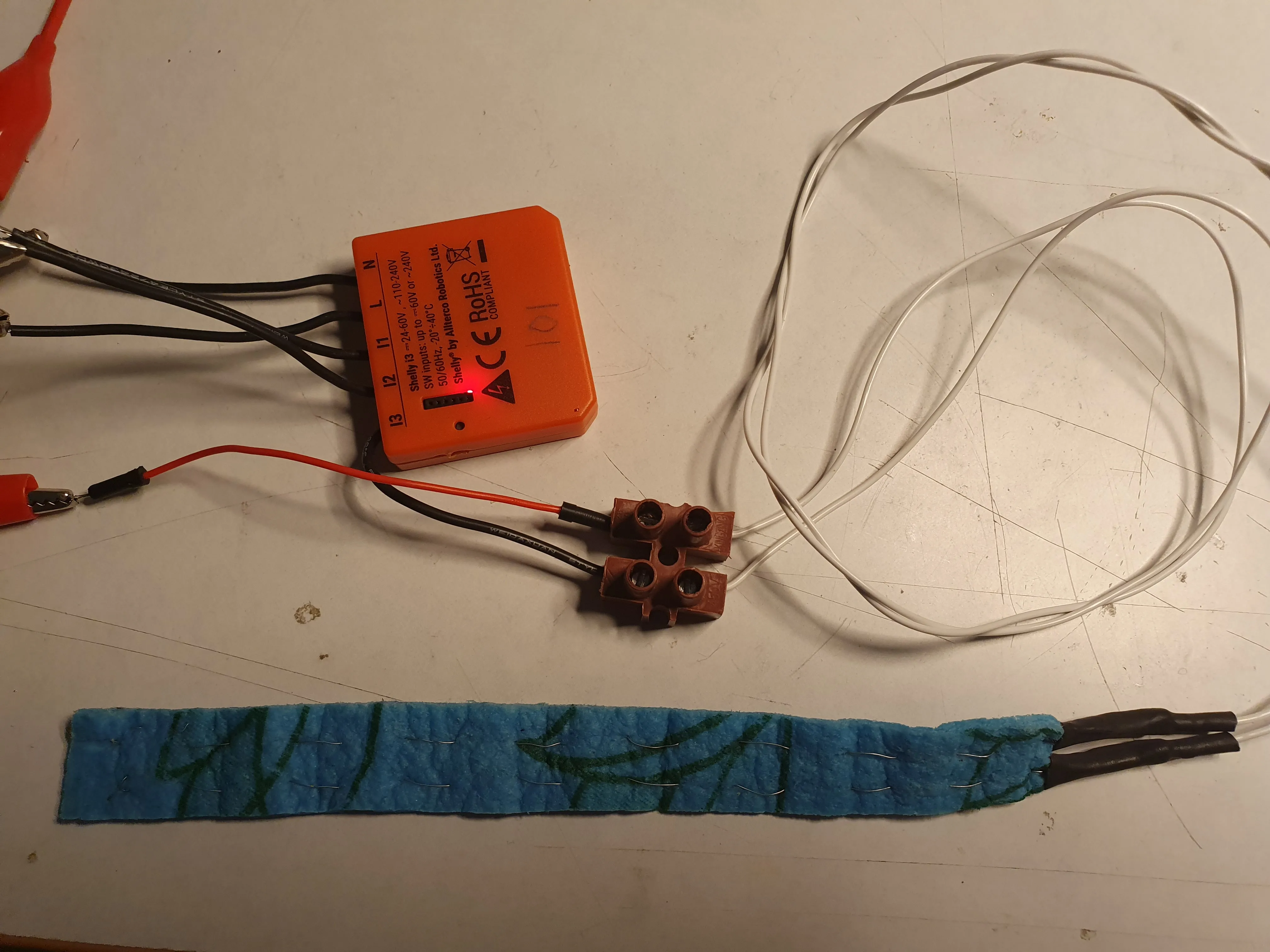 An orange Shelly i3 wired to a strip of blue sponge cloth with two bare wires stitched through it as the sensing element